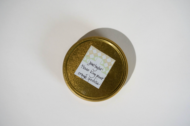 A gold metal tin with a white and green patterned label reading June Taylor Pluot Pastille by Broc Cellars sits on a white surface, casting a rightward shadow. The tin’s design recalls classic June Taylor Orange Pastille packaging.