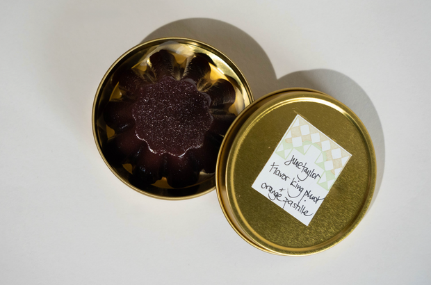 A small round gold tin with a patterned label reading Broc Cellars June Taylor Pluot Pastille sits open on a light surface, revealing a dark purple, flower-shaped solid inside reminiscent of the Flavor King Pluot.
