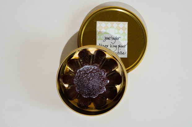 A round gold tin from Broc Cellars, labeled on the lid, holds a single dark, flower-shaped June Taylor Pluot Pastille dusted with sugar, seen from above on a plain white surface.