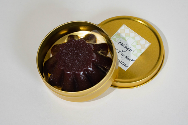A round gold tin from Broc Cellars, labeled "June Taylor Pluot Pastille," sits open on a white background, revealing a dark red, fluted, flower-shaped pastille inside. The lid features a small handwritten label.