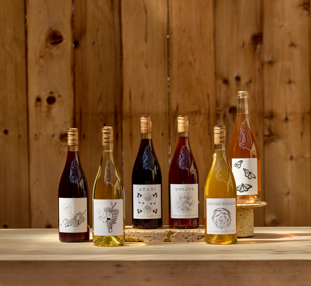 Six bottles of Broc Cellars 2026 SUMMER RELEASE-CLASSIC wines, with illustrated labels, are arranged on corks atop a wooden table against a fence. The set features red, rosé, and white varieties with gold caps.