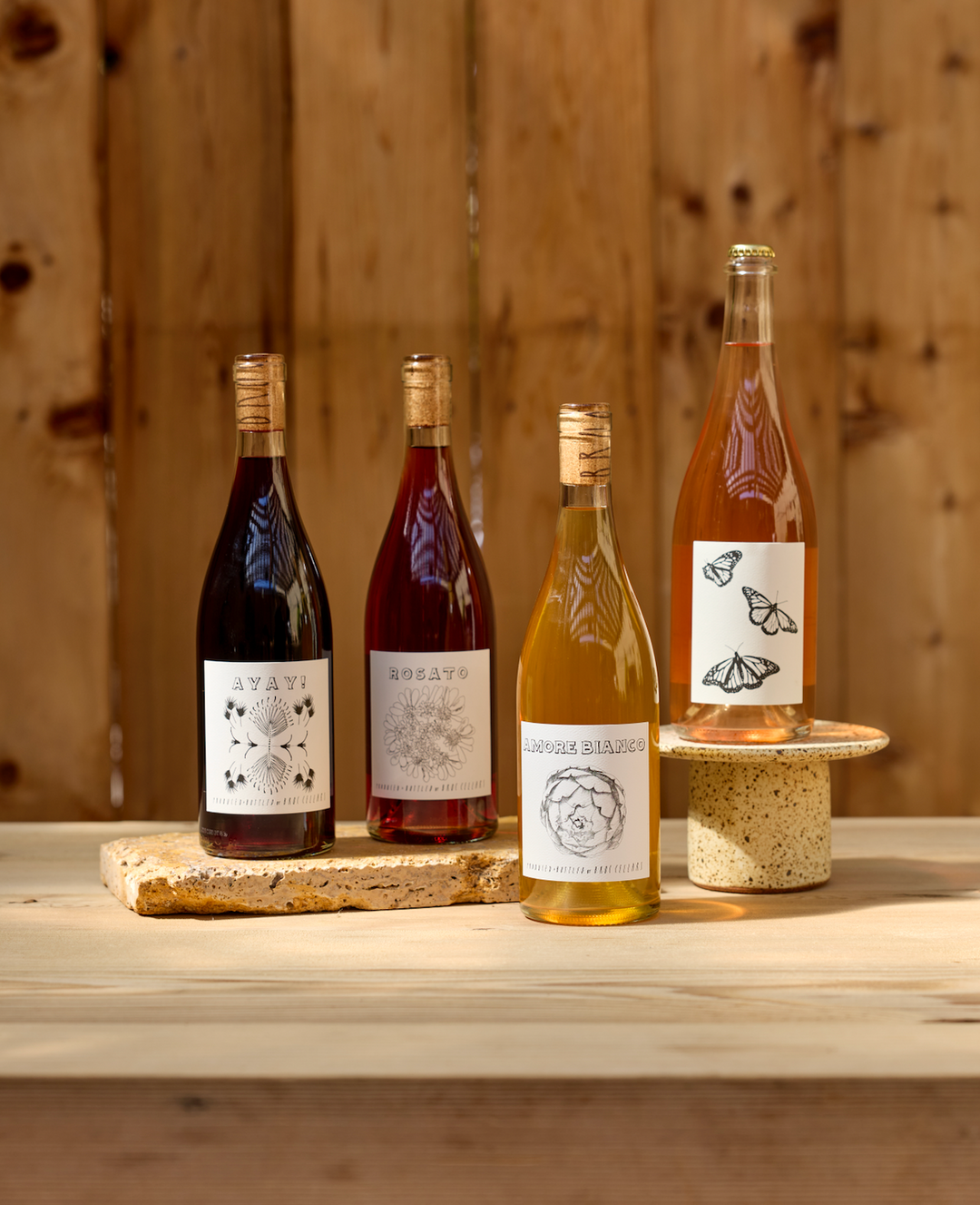 Four bottles of Broc Cellars 2026 SUMMER RELEASE - LITE wine, some with illustrated labels, stand on a wooden table before a wood background; one bottle is elevated on a small round pedestal.