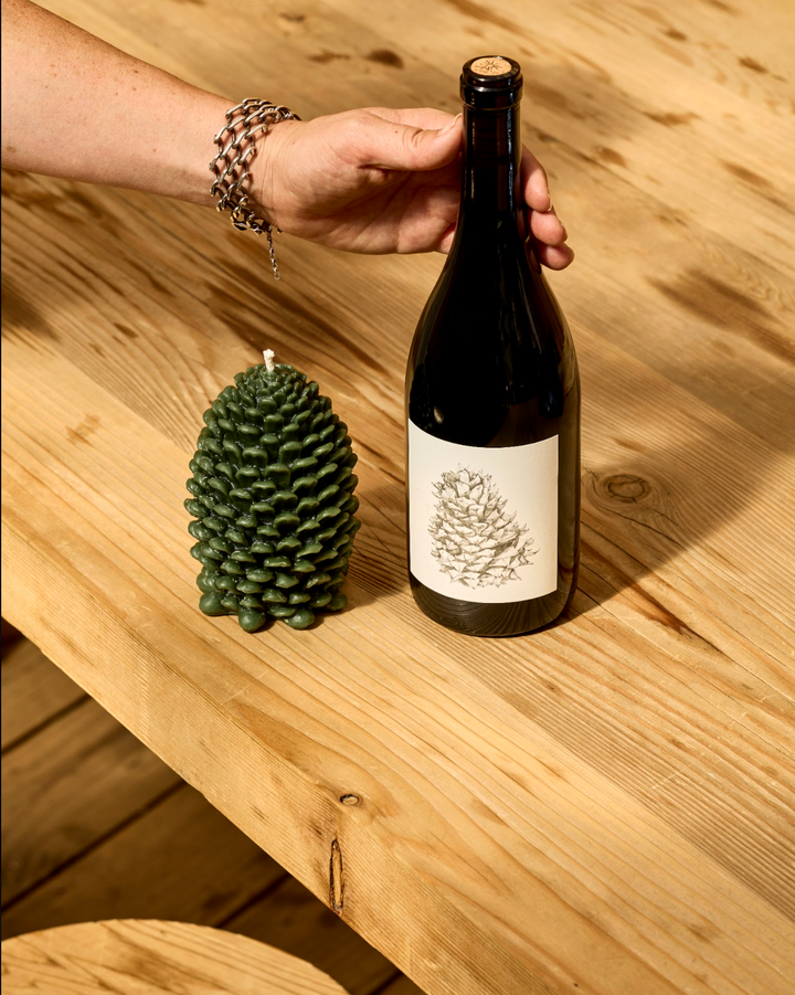 A person’s hand holds a bottle of Broc Cellars Lagrein &amp; Pine Candle wine next to a dark green pinecone-shaped beeswax candle on a wooden table.