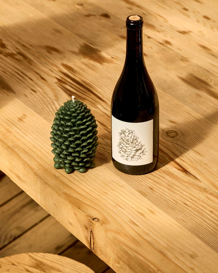 A dark green pinecone-shaped beeswax candle from the Broc Cellars Lagrein &amp; Pine Candle set sits next to a bottle of Lagrein wine featuring a pinecone illustration on the label, all placed on a wooden table.