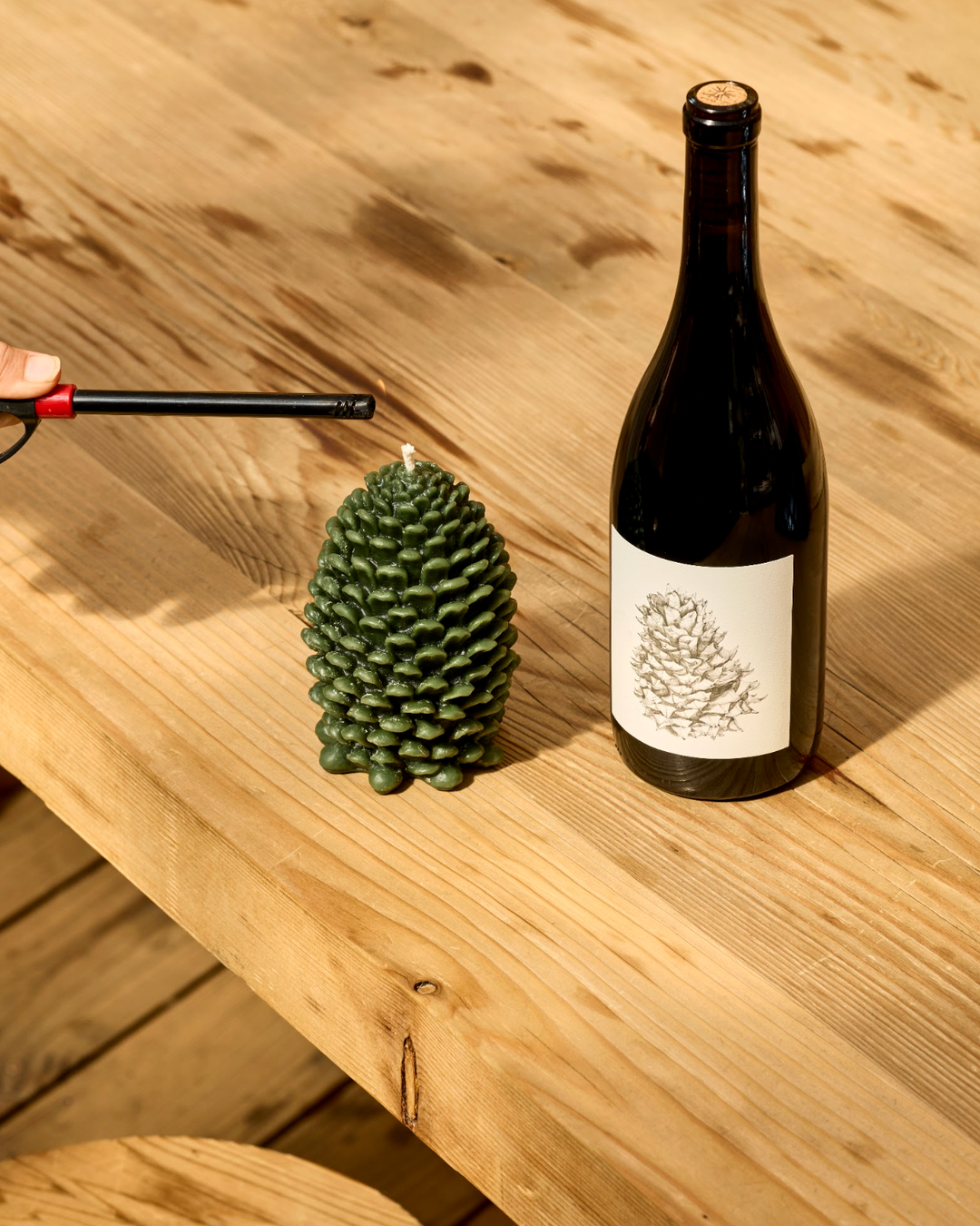 A hand uses a lighter to ignite a green pinecone-shaped Broc Cellars Lagrein &amp; Pine Candle next to a bottle of Lagrein wine featuring a matching pinecone illustration on the label.