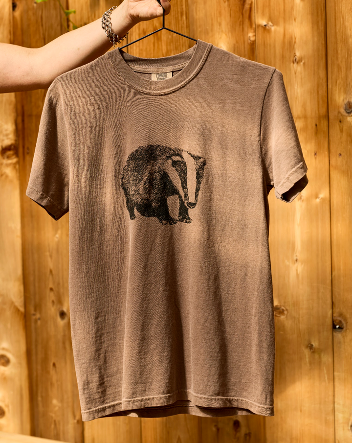 Brown short-sleeve Comfort Colors unisex Badger T-Shirt with a black badger illustration on the chest, held up on a hanger.