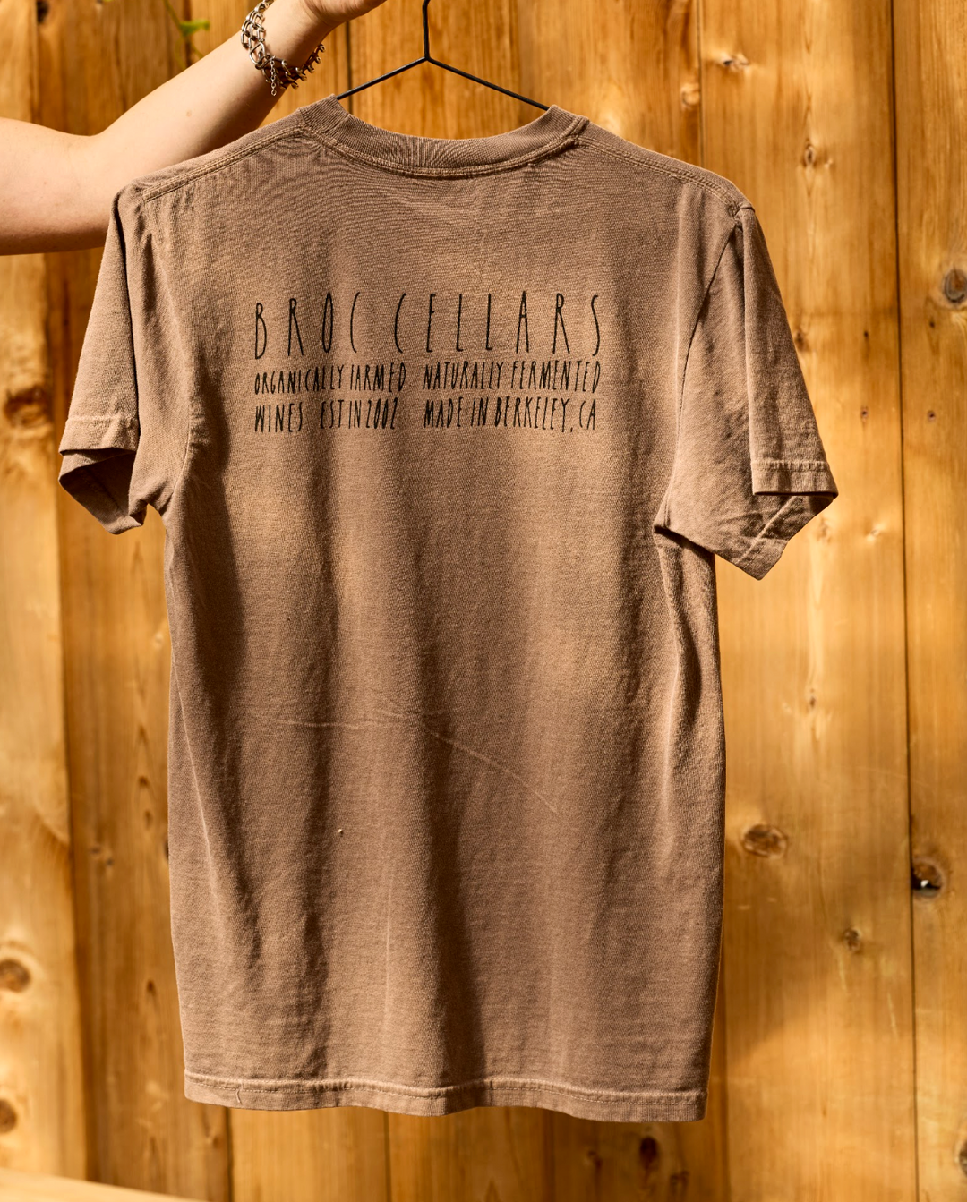 Brown unisex Badger T-Shirt by Broc Cellars hanging on a hanger, featuring text that reads Broc Cellars, Organically Farmed, Naturally Fermented, Wines Est in 2002, Made in Berkeley, CA.