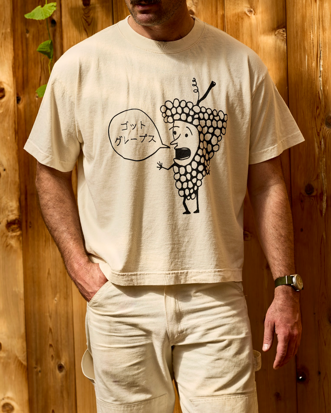 A person wearing a cream-colored, 100% cotton Got Grapes T-Shirt by Broc Cellars, featuring a cartoon grape cluster graphic with a speech bubble containing Japanese text.