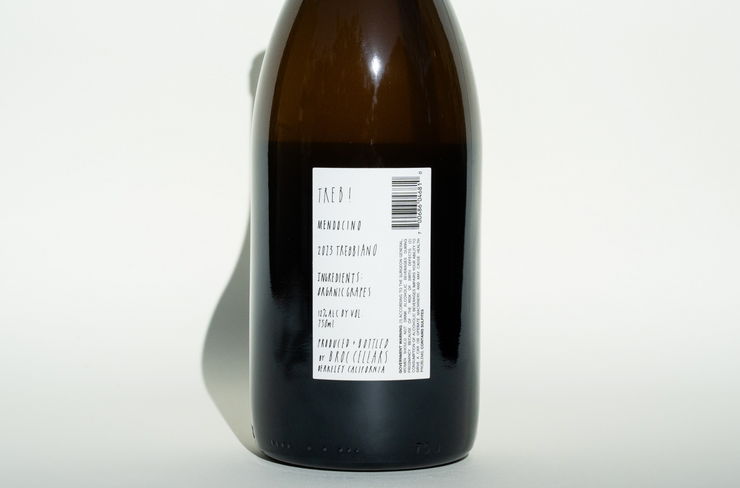 A dark glass bottle features a white label with vertically handwritten text, stating Broc Cellars and 2023 Treb! Shadows on the white surface suggest strong light from the left, enhancing the allure of this California wine.