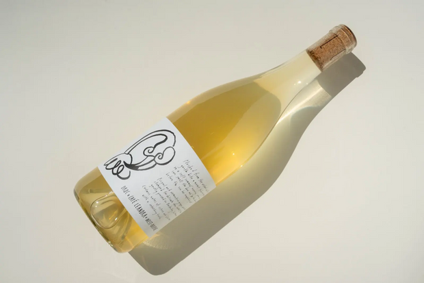 A bottle of Broc Cellars Broc x Café Leandra x West-Bourne white wine rests on its side, label featuring black line art. Crafted from Picpoul and Vermentino grapes, it offers citrus and blossom notes.
