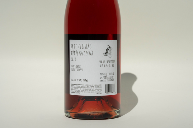 A close-up of a bottle of Broc Cellars 2024 Rosato with a pink-red hue, focusing on the back label with vintage, producer, and Fox Hill Vineyard origin. A shadow falls across the white surface. Certified Organic.