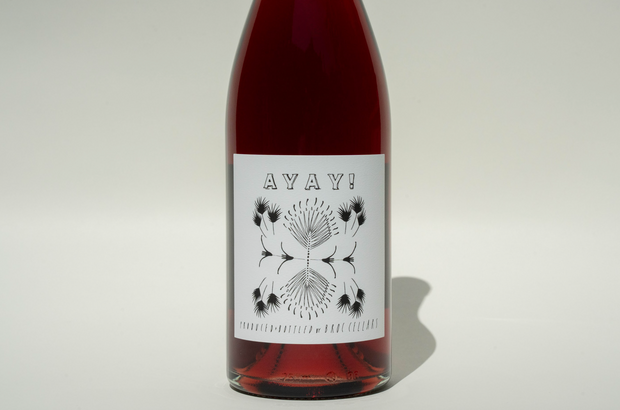 A bottle of 2024 Ayay! wine from Broc Cellars with a white label featuring AYAY! in bold above a symmetrical black and white botanical illustration, set against a plain, light background.