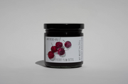 A glass jar with a black lid contains June Taylor x Fox Hill Wild Cherry Butter by Broc Cellars. The white label features an illustration of five red plums, listing the contents and net weight, set against a plain light background.