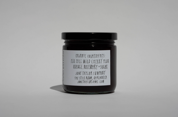A small glass jar with a black lid and white label lists wild cherry plum, orange, rosemary, and sugar. Broc Cellars June Taylor x Fox Hill Wild Cherry Butter is a hand-made preserve with the website also shown on the label.