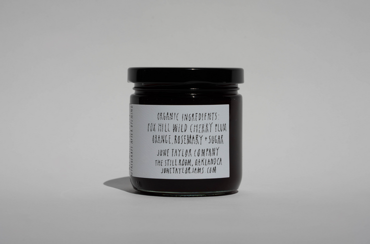 A small glass jar with a black lid and white label lists wild cherry plum, orange, rosemary, and sugar. Broc Cellars June Taylor x Fox Hill Wild Cherry Butter is a hand-made preserve with the website also shown on the label.