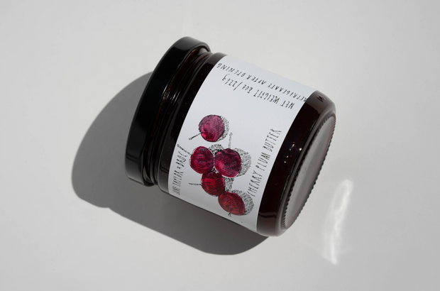 A dark glass jar of June Taylor x Fox Hill Wild Cherry Butter by Broc Cellars, with a white label showing red cherries and black text, lies on its side on a white surface. Some label details are partially visible around the edge.