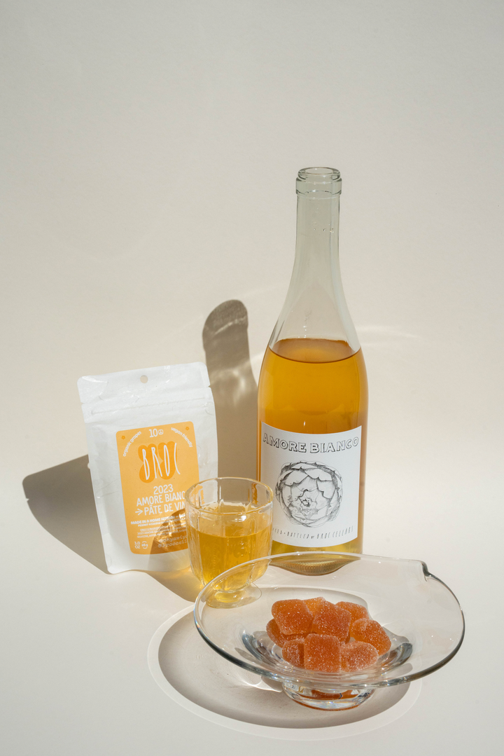 A bottle of Broc Cellars Amore Bianco + Yami Yami Pâte de Vin, a filled glass, a pouch, and orange candies are stylishly arranged on a white surface.