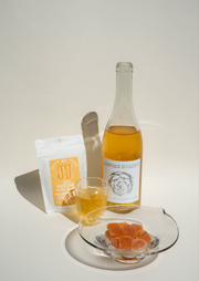 A bottle of Broc Cellars Amore Bianco + Yami Yami Pâte de Vin, a filled glass, a pouch, and orange candies are stylishly arranged on a white surface.