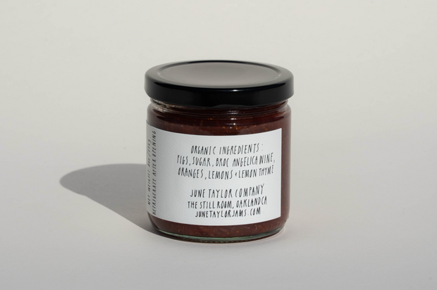 A glass jar with a black lid contains June Taylor Mission Fig + Angelica Jam by Broc Cellars. The label lists figs, sugar, dark muscatel wine, oranges, lemons, and lemon thyme. This flavorful preserve is made in Oakland, CA.