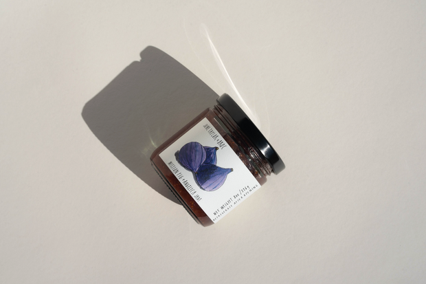 A small glass jar of Broc Cellars June Taylor Mission Fig + Angelica Jam with a black lid rests on a light surface. The label displays a blue-purple flower and text, while the jar casts a distinct shadow to the left.