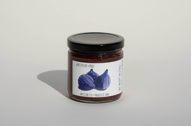 A glass jar with a black lid holds Broc Cellars' June Taylor Mission Fig + Angelica Jam. The label shows two figs and the product name, with smaller text on the side.