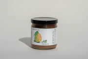 A glass jar with a black lid contains Broc Cellars’ June Taylor Fox Hill Pear, Lemon Geranium, and Rosemary Preserve. The white label features a watercolor pear, green leaves, product name, and opening instructions, emphasizing its artisanal quality.