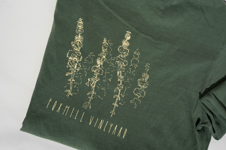 The Broc Cellars Fox Hill Organic Long Sleeve Shirt is made in the USA from 100% cotton. This green unisex shirt features a light yellow line drawing of tall plants with "FOXHILL VINEYARD" below the illustration for a comfy fit.