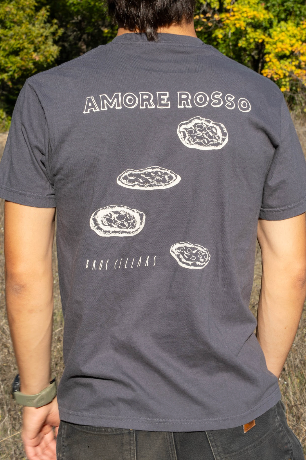 Outdoors, a person wears a dark Broc Amore Rosso T-Shirt by Broc Cellars, featuring the Amore Rosso logo and three pizzas above "HAND CULLED," with trees and dry grass in the background.