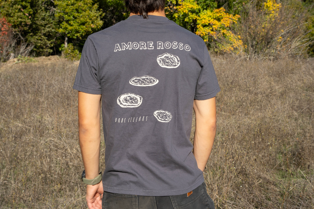 Outdoors, a person wears a dark Broc Amore Rosso T-Shirt by Broc Cellars, featuring the Amore Rosso logo and three pizzas above "HAND CULLED," with trees and dry grass in the background.