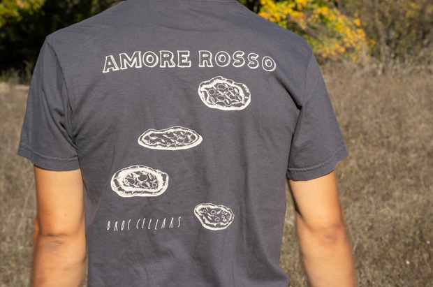 Outdoors, someone wears a dark gray unisex Broc Amore Rosso T-Shirt by Broc Cellars, featuring the logo, "BROC CELLARS," and three oyster graphics on the back. Dry grass and trees recall the rustic Broc Pocket Pizzeria vibe.