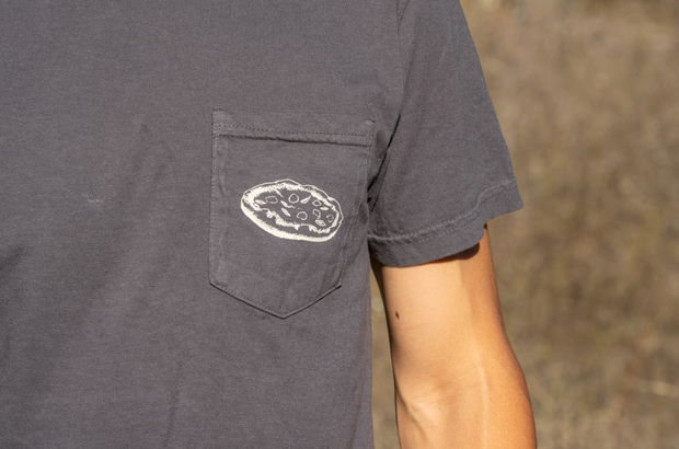 A person wears the Broc Cellars Broc Amore Rosso T-Shirt, a dark gray unisex tee with a chest pocket and circular Amore Rosso logo patch. They stand outdoors against a blurred brown background.