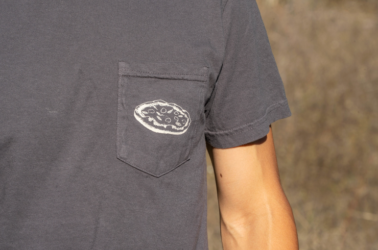 A person wears the Broc Cellars Broc Amore Rosso T-Shirt, a dark gray unisex tee with a chest pocket and circular Amore Rosso logo patch. They stand outdoors against a blurred brown background.