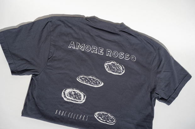 A dark gray unisex Broc Amore Rosso T-Shirt by Broc Cellars lies flat, showing the back with the Amore Rosso logo above four illustrated oyster shells and “DNO CELLARS” printed near the bottom.
