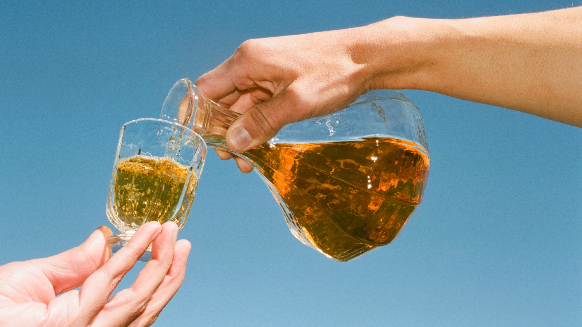 A hand pours golden 2024 Amore Bianco from Broc Cellars into a small glass held under a clear blue sky—refreshment is moments away.