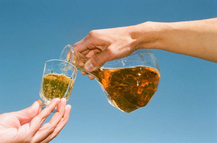 A hand pours golden 2024 Amore Bianco from Broc Cellars into a small glass held under a clear blue sky—refreshment is moments away.