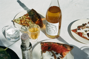 Three plates with pizza slices, a bottle of Broc Cellars 2024 Amore Bianco, a wine glass, and a glass of water sit on a white tablecloth in natural light. A partially visible bottle hints at more Broc Cellars wines to come.