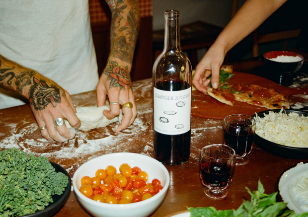 Two people prepare food on a floured table with cherry tomatoes, greens, grated cheese, and a bottle of Broc Cellars 2023 Amore Rosso. One rolls dough as the other holds a slice of pizza, creating a perfect pairing experience.