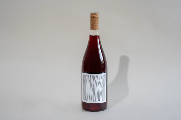 A bottle of Broc Cellars 2025 Nouveau with a cork closure sits on a plain, light background. The white label displays abstract, vertical black lines without readable text, and the bottle casts a soft shadow to the right.