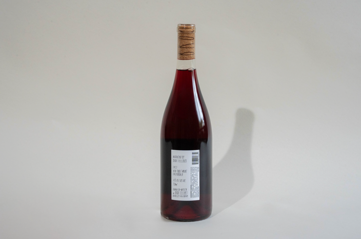 A clear glass bottle of dark red liquid stands upright against a light gray background, sealed with a cork. The white label reads "2025 Nouveau" from Broc Cellars and shows printed text and a barcode.