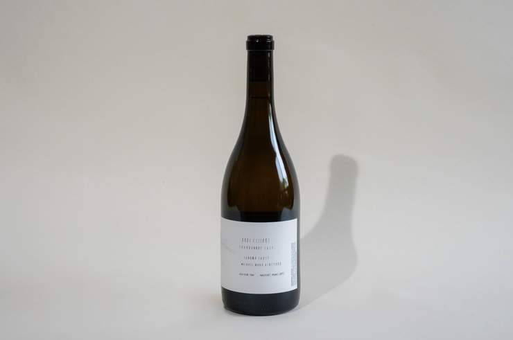 A single, unopened bottle of Broc Cellars 2024 Michael Mara Chardonnay with a white label and black cap stands on a plain, light gray surface. Its label hints "Coming Soon" in small text as the bottle's shadow stretches rightward.