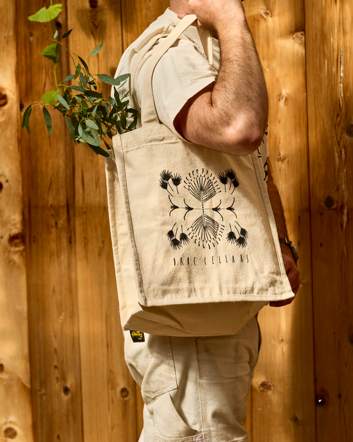 A person carries a tan canvas Broc 4 Bottle Canvas Bag containing a leafy green plant, with a black botanical print and text visible on the side.