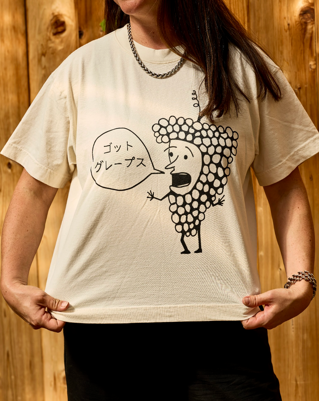 A person wears a cream Got Grapes T-Shirt, an oversized garment featuring a black line-drawing of a cartoon grape character with a speech bubble containing Japanese text.