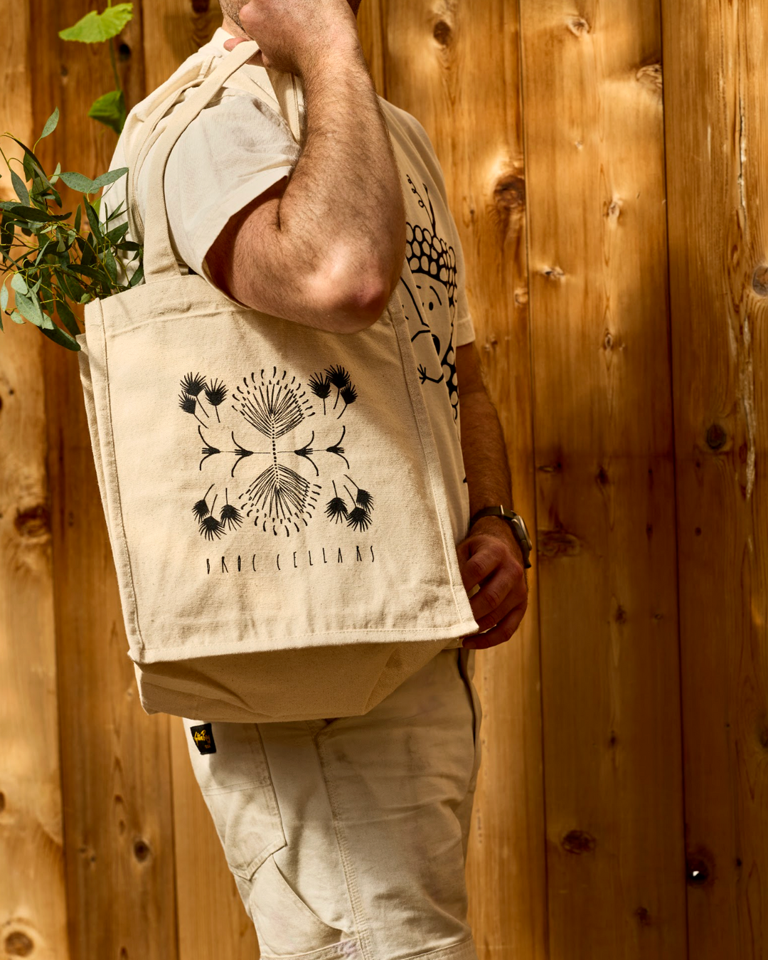 A person carries a tan canvas Broc 4 Bottle Canvas Bag featuring a black botanical illustration and text on their shoulder, with greenery poking out of the top.