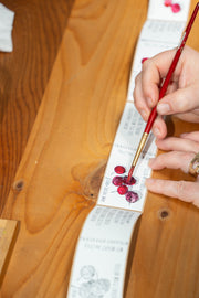 A person uses a paintbrush to add color to Broc Cellars June Taylor x Fox Hill Wild Cherry Butter labels, each featuring berry art. Several customized labels are lined up on a wooden table, ready for the handmade jam jars.