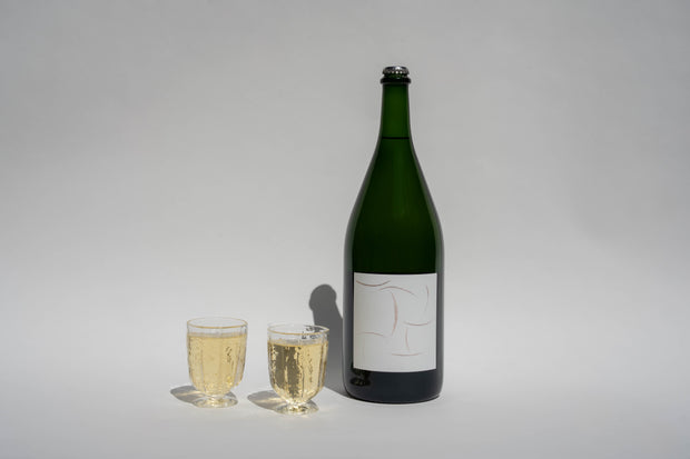 A green bottle of Broc Cellars 2024 Sparkling Chenin Magnum with a blank white label stands beside two filled Broc glasses on a plain white background.