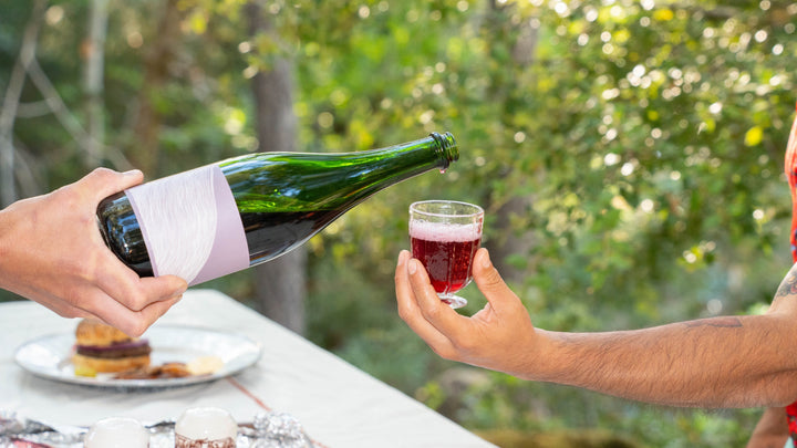 At an outdoor table with desserts in daylight, one person pours Broc Cellars 2024 Aqua Vino, a dark red, low-alcohol sparkling wine from a green bottle, into a small glass held by another.