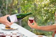 At an outdoor table with desserts in daylight, one person pours Broc Cellars 2024 Aqua Vino, a dark red, low-alcohol sparkling wine from a green bottle, into a small glass held by another.
