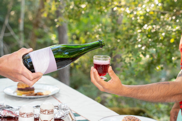 At an outdoor table with desserts in daylight, one person pours Broc Cellars 2024 Aqua Vino, a dark red, low-alcohol sparkling wine from a green bottle, into a small glass held by another.