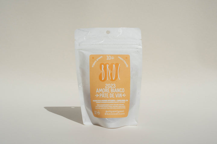 A white stand-up pouch with an orange label reading "Amore Bianco + Yami Yami Pâte de Vin" by Broc Cellars stands upright on a neutral background, suggesting a skin-contact white wine, with extra information beneath.