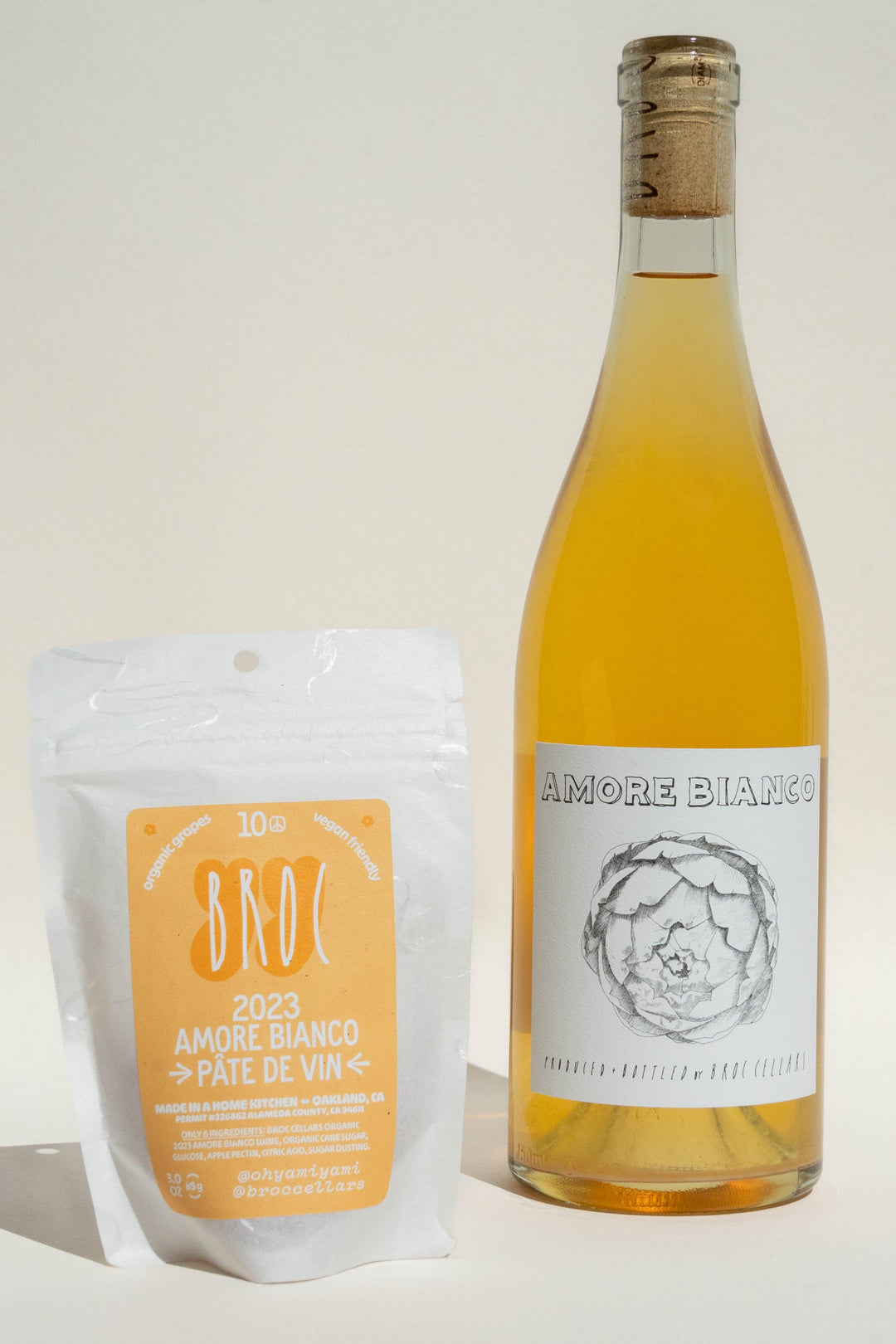 A bottle of Broc Cellars Amore Bianco + Yami Yami Pâte de Vin stands beside a clear pouch labeled “2023 Amore Bianco Pâté de Vin” against a light background.